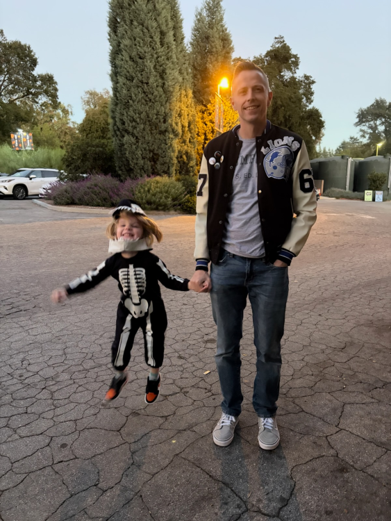 A full-body shot of Patrick, dressed in a black and white letterman jacket and jeans as Axel Foley from Beverly Hills Cop. He stands next to a smiling young child in a black and white skeleton costume and hat. The child is holding Patrick's hand while leaping high into the air. They are standing on a paved surface outdoors at dusk.