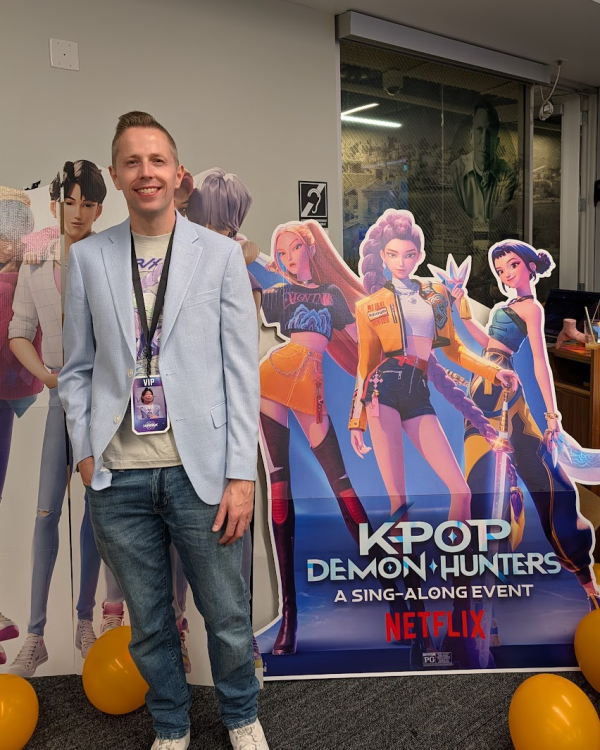 Patrick wearing a Halloween costume of Bobby from K-Pop Demon Hunters, smiles as he stands next to a large promotional cutout for the Netflix movie, K-Pop Demon Hunters: A Sing-Along Event. He is dressed in a light blue blazer over a light gray t-shirt, and blue jeans.