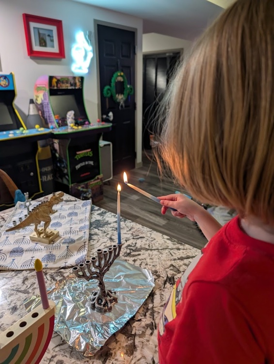 A young child named Patrick, with chin-length blonde hair and wearing a red shirt, uses a slender blue-and-white candle to light the first candle of a silver tree-shaped menorah. The menorah sits on a piece of aluminum foil on a granite countertop. In the background, a cozy room features arcade games, including a Teenage Mutant Ninja Turtles machine, a decorative gold dinosaur menorah, and a lit neon sign on the wall.